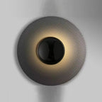 Larleon woven mesh disc halo wall sconce — dark textured disc with glossy black center sphere and warm radial LED gradient on grey wall